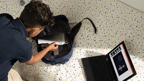 A boy is looking into his bag with his laptop on the bed