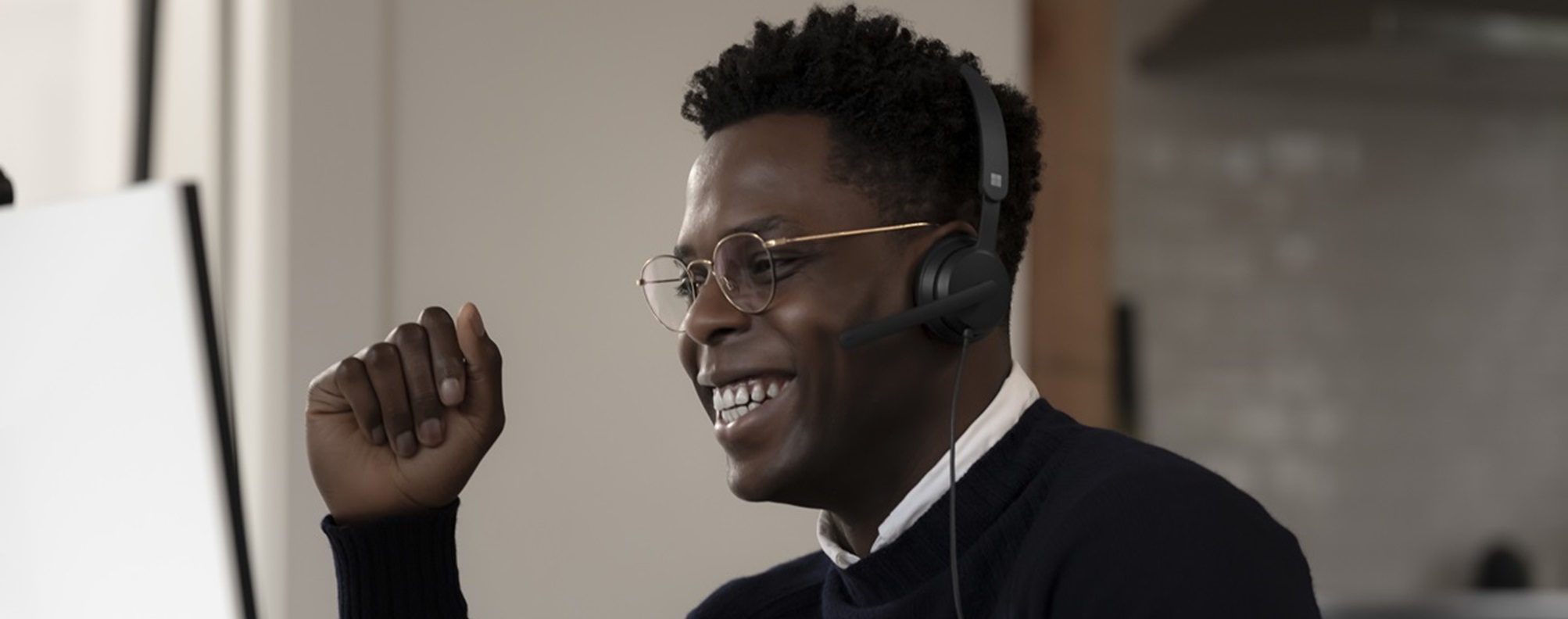 Young man wearing glasses and a headset smiling at a computer monitor with a mounted webcam