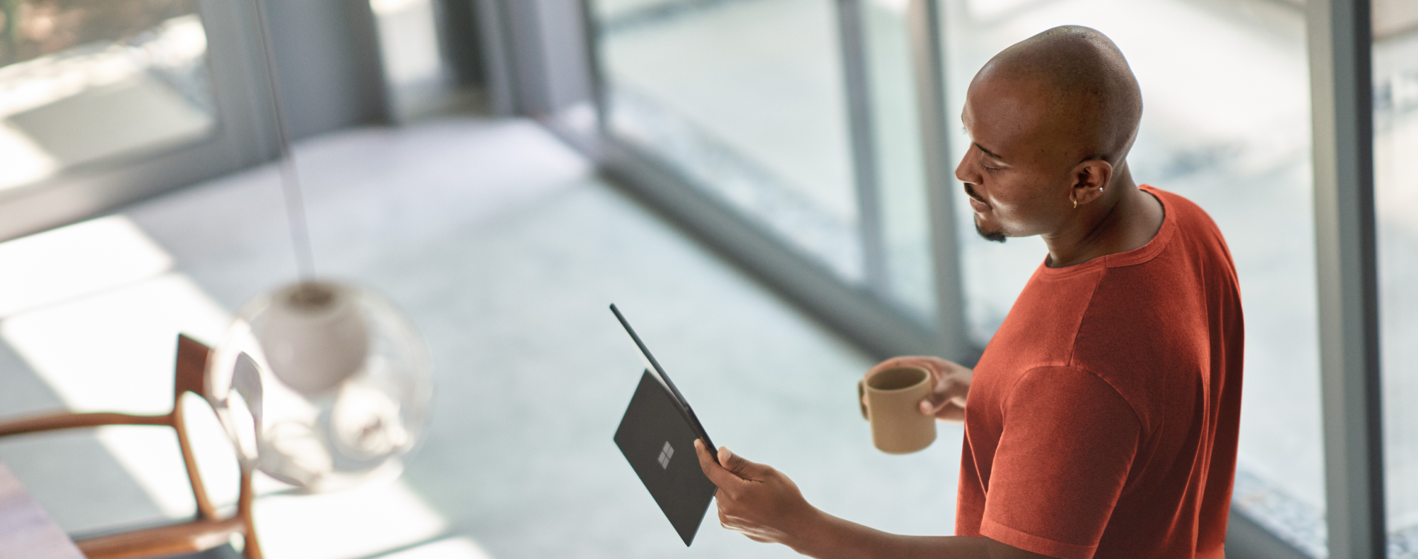  Person standing indoors near large windows, holding a tablet in one hand and a coffee mug in the other, with modern furniture and natural light in the background.