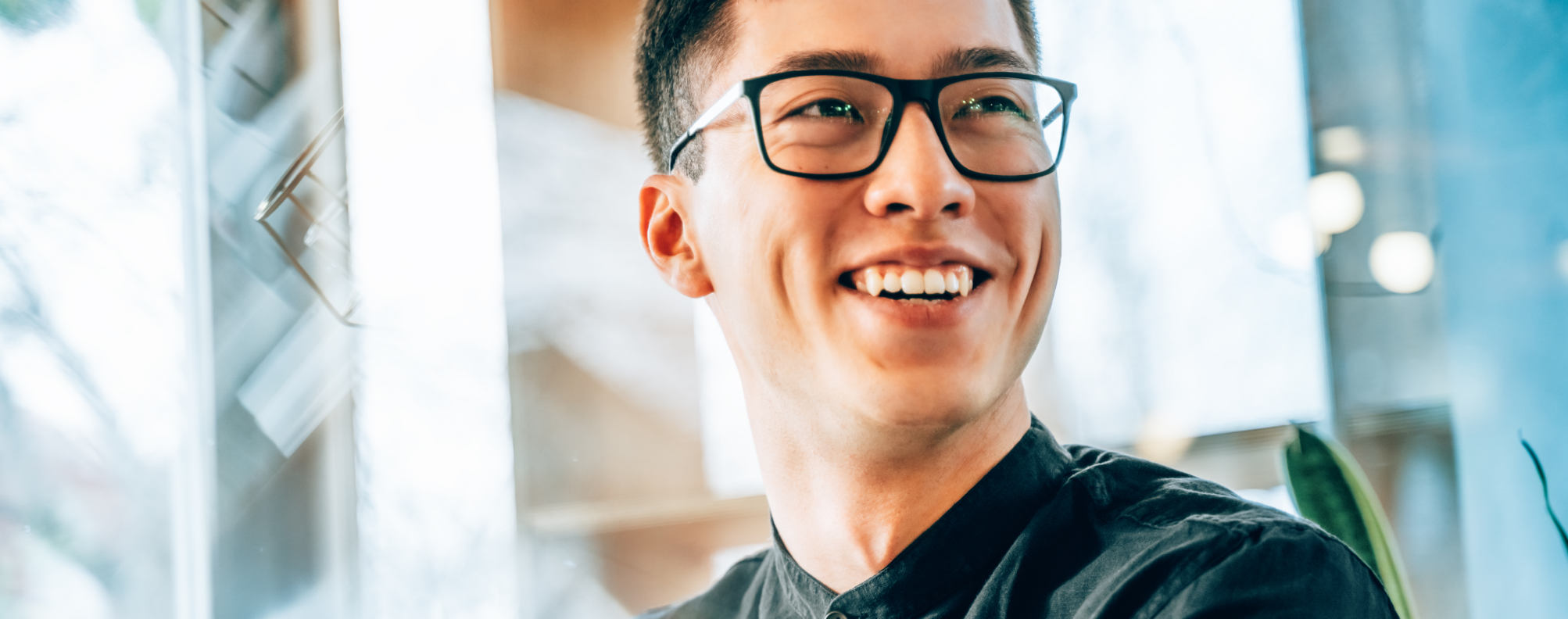 Smiling professional wearing glasses, seated in a bright modern office, with natural light and greenery in the background, conveying optimism and innovation.