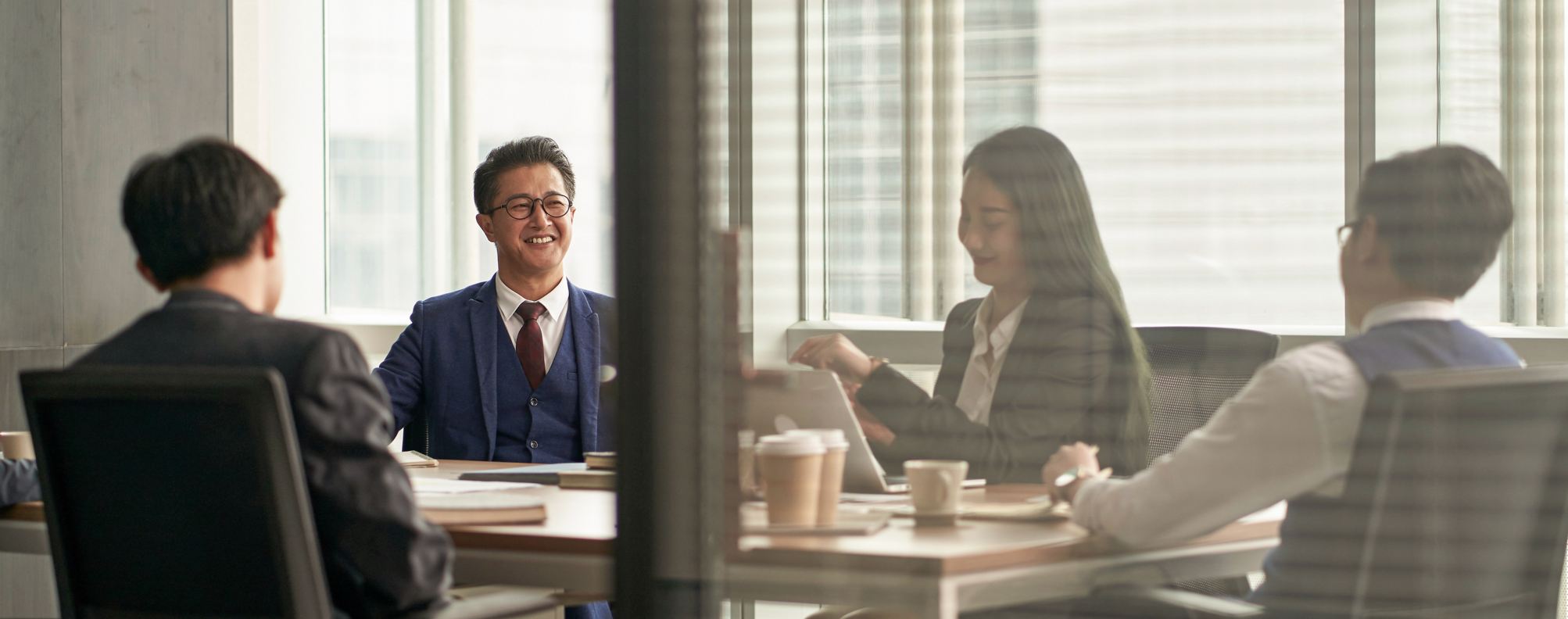  Four people in business attire seated around a conference table with notebooks, coffee cups, and documents in a modern office setting.