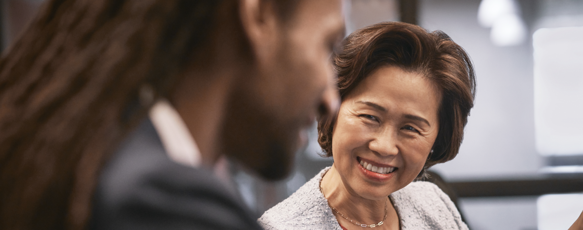 Two professionals seated in conversation at a modern office table, with focus on one person’s styled hair, necklace, and textured jacket against a softly lit background.