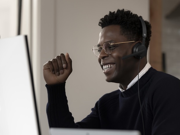 Young man wearing glasses and a headset smiling at a computer monitor with a mounted webcam