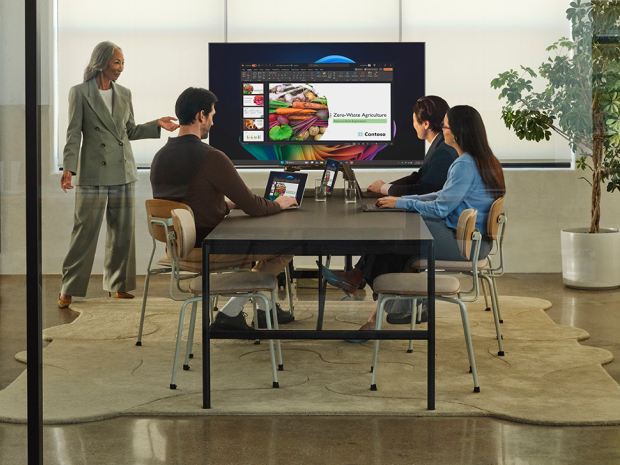 Four people collaborate in a modern office meeting room using laptops and a large digital presentation screen.