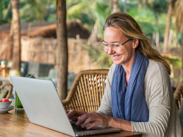 Person working on a laptop at an outdoor wooden table with tropical plants and thatched structures in the background.