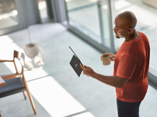 Person standing indoors near large windows, holding a tablet in one hand and a coffee mug in the other, with modern furniture and natural light in the background.