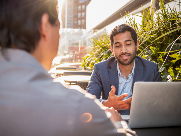 Two people at an outdoor cafe having a conversation over a laptop.