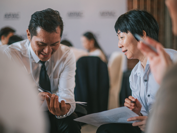 Two people in formal attire sitting close together, holding papers and engaged in a discussion during a professional event.