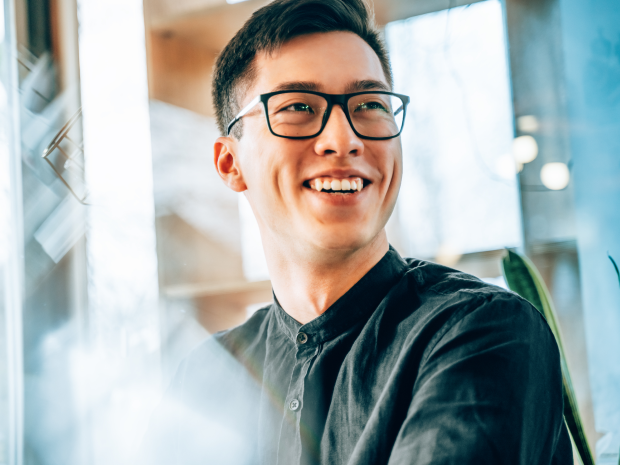 Smiling professional wearing glasses, seated in a bright modern office, with natural light and greenery in the background, conveying optimism and innovation.