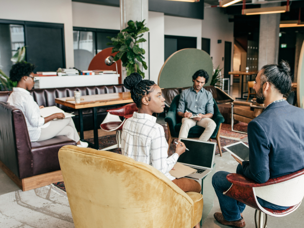 A group of businesspeople collaborating in a work lounge while using laptops and tablets.