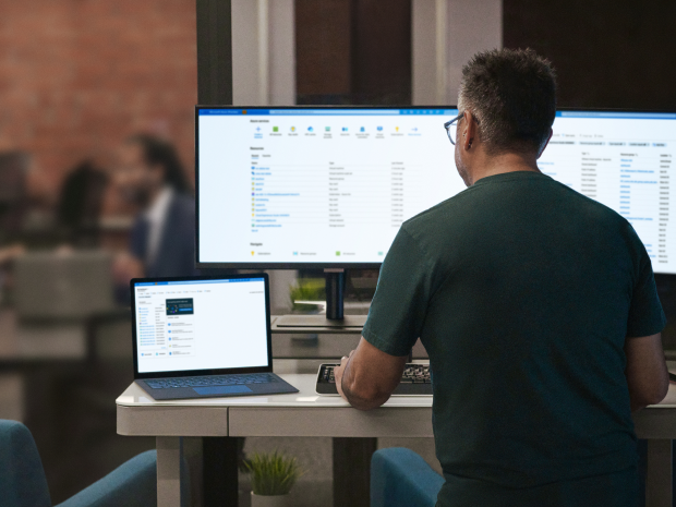 Person working at a desk with three monitors displaying file management software, viewed from behind. The background shows a blurred meeting room with people seated at a table.