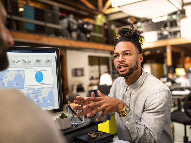 Person discussing data insights displayed on a monitor showing analytics charts and global activity maps in a modern office.