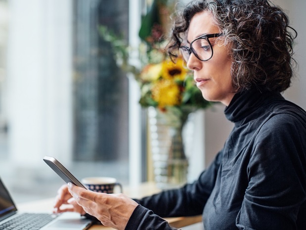 Woman sitting at a desk with her laptop and looking at her phone in her hand.