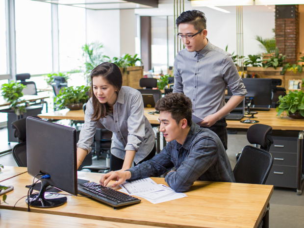 Three people collaborate at a computer in a modern, open-plan office with natural light and green plants.