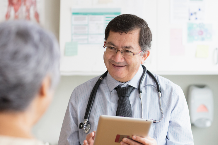 A smiling doctor holding a tablet and talking to a patient.
