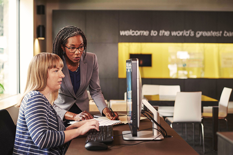 Two women at a desk look at a computer screen One of the women is standing while the other is seated and using a keyboard