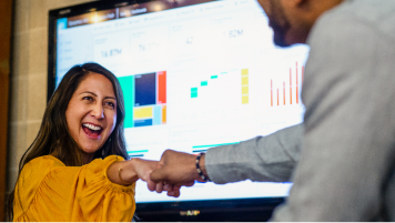 Two people fist bumping near a screen with data charts.